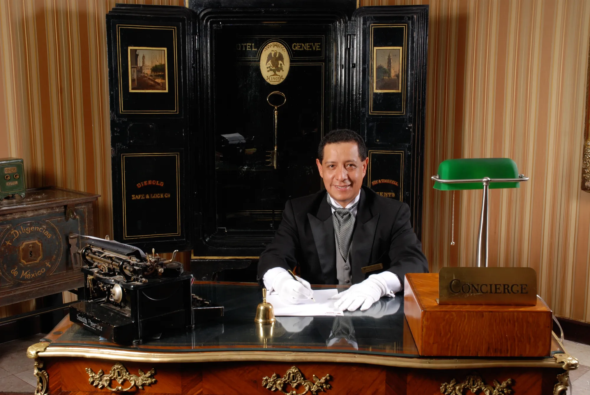 Concierge sonriendo detr&aacute;s de un escritorio con una l&aacute;mpara verde y una m&aacute;quina de escribir antigua. Fondo con caja fuerte.