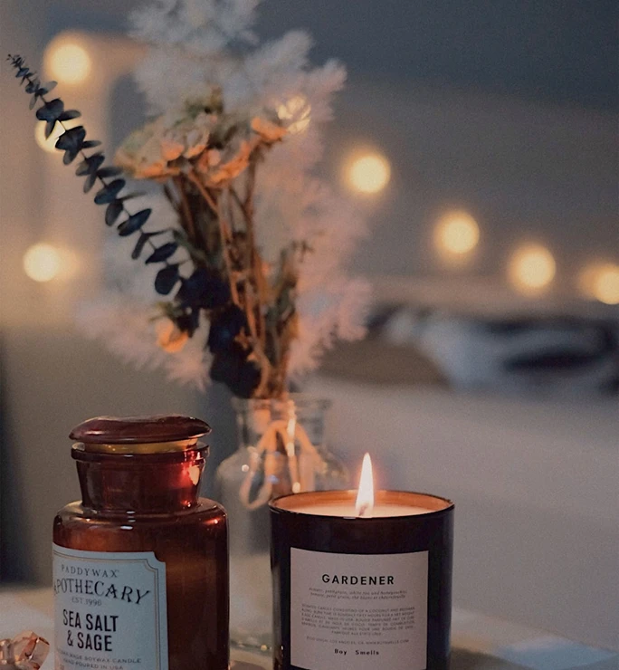 Vela encendida junto a un frasco marr&oacute;n y un jarr&oacute;n con flores secas. Luces difusas de fondo crean ambiente acogedor.