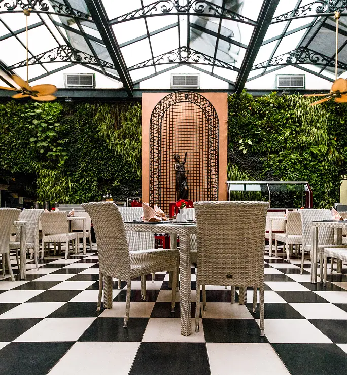 Restaurante elegante con techo acristalado, suelo ajedrezado, muebles de mimbre y pared verde con estatua decorativa.