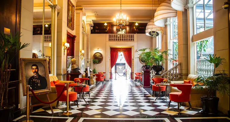 Sal&oacute;n elegante con suelo ajedrezado, cuadros, candelabros, sillas rojas y plantas, iluminado con luz natural.