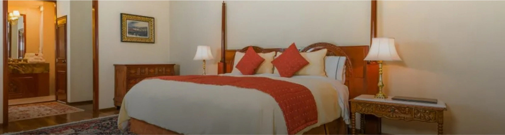 Habitaci&oacute;n con cama grande, edred&oacute;n rojo y l&aacute;mparas a los lados. Muebles de madera y decoraci&oacute;n cl&aacute;sica.