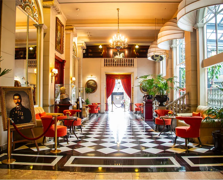 Sal&oacute;n elegante con pisos de damero, candelabro, retrato y sillas naranjas. Puerta roja al fondo, plantas y luz natural.