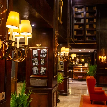 Vest&iacute;bulo elegante con iluminaci&oacute;n c&aacute;lida, sillones rojos, plantas y estanter&iacute;as llenas de libros.