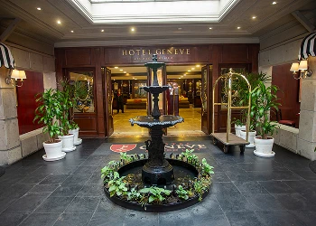 Entrada del Hotel Geneve con una fuente central rodeada de plantas y un carrito de equipaje a la derecha.