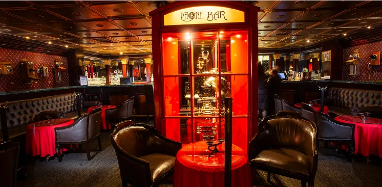 Bar tem&aacute;tico con cabina telef&oacute;nica roja y decoraci&oacute;n elegante, con sof&aacute;s oscuros y mesas rojas alrededor.