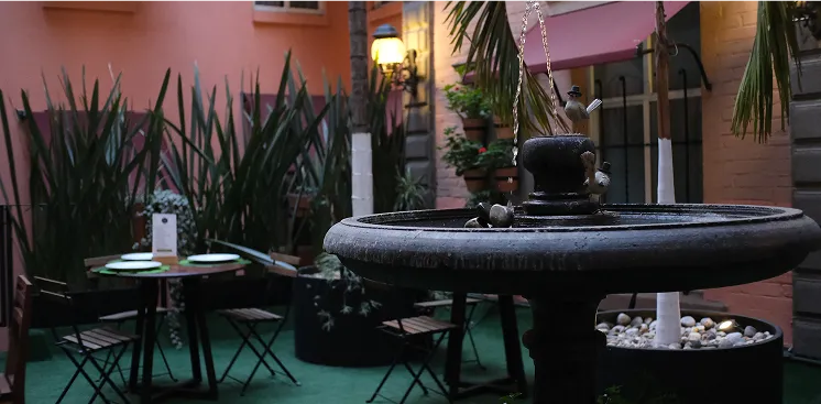 Un patio con fuente central, mesas y sillas rodeadas de plantas, bajo una luz c&aacute;lida y paredes rosadas.