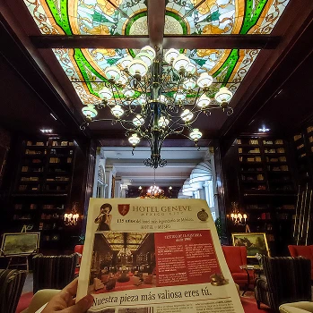 Sala elegante con techo de vitrales y candelabro. Persona sostiene folleto del Hotel Geneve. Sof&aacute;s y libros alrededor.