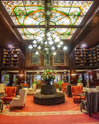 Lobby elegante con candelabro, vitral en el techo, sillones alrededor de una mesa central con flores, estanter&iacute;as de libros.