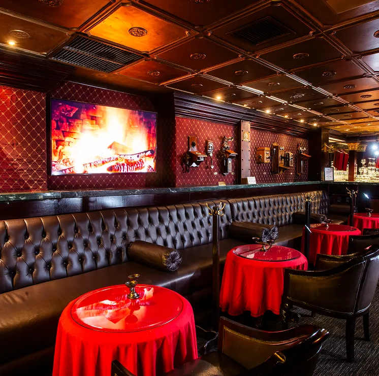 Bar elegante con mesas redondas cubiertas por manteles rojos, paredes acolchadas, y pantalla con chimenea encendida.