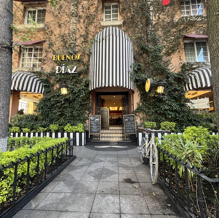 Entrada de un caf&eacute; con toldo a rayas, plantas trepadoras, ventanas y una bicicleta estacionada enfrente.