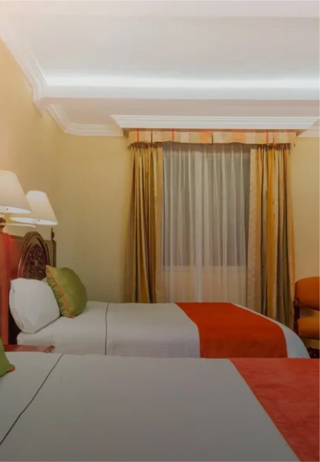 Habitaci&oacute;n de hotel con dos camas individuales, con cojines verdes y colchas anaranjadas; cortinas doradas.