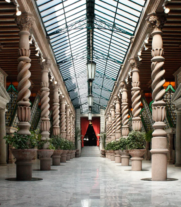 Elegant corridor with spiral columns, glass roof, and potted plants, leading to red curtains at the end.
