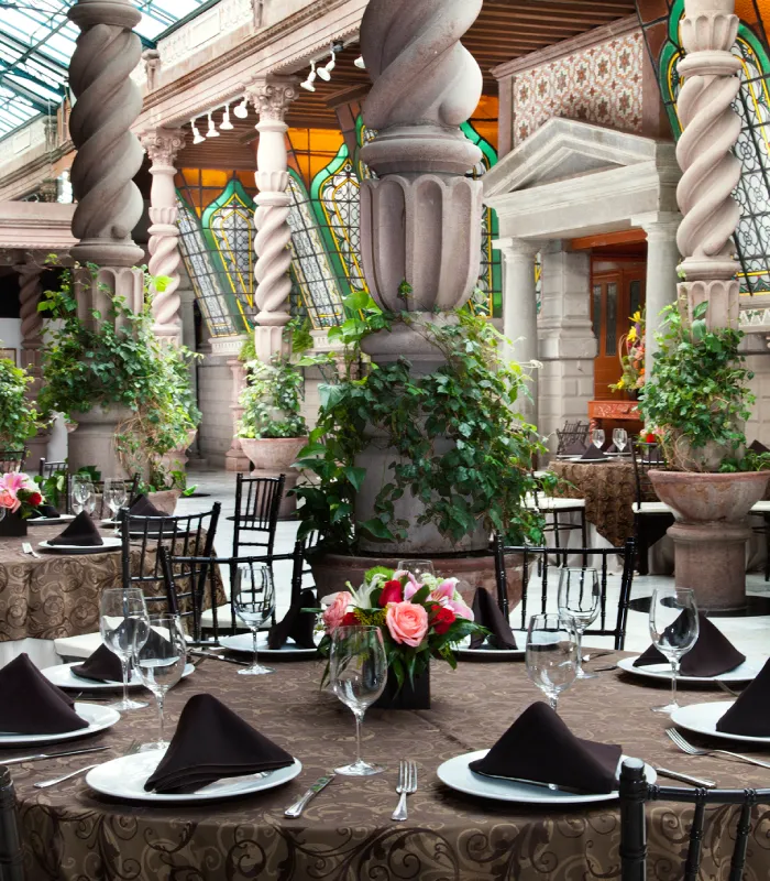 Decorated dining area with ornate columns, plants, tables set with plates and glasses, and stained-glass windows.