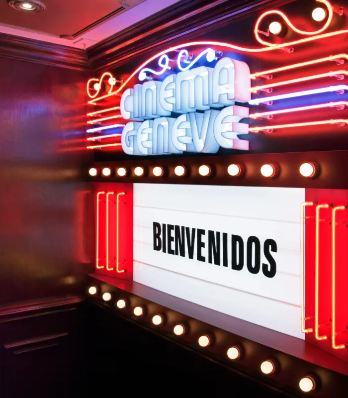 Cinema Gen&egrave;ve neon sign with "BIENVENIDOS" on a lit marquee, surrounded by colorful lights.