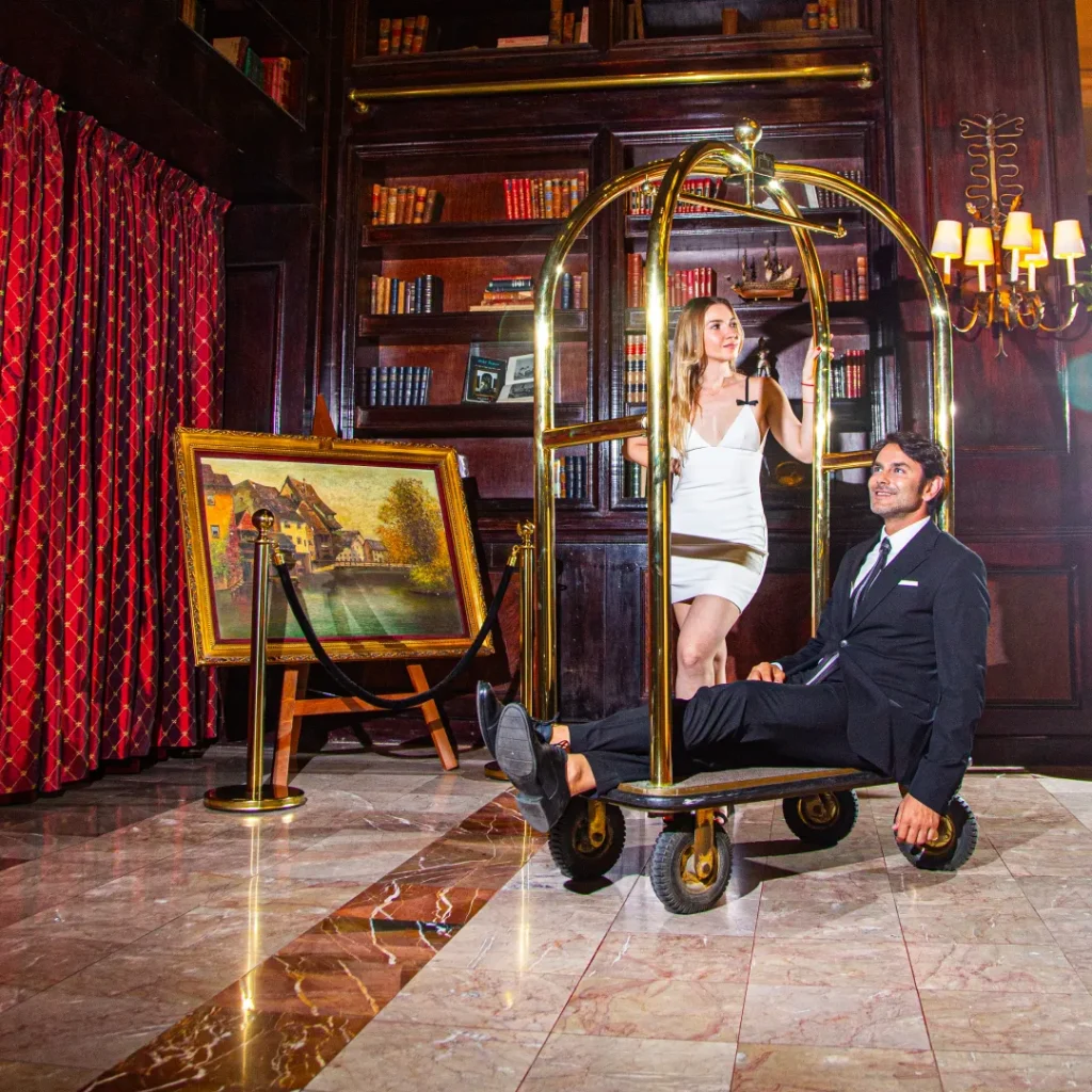 Personas posan en un carrito de equipaje en una biblioteca elegante, con pintura y luces al fondo.