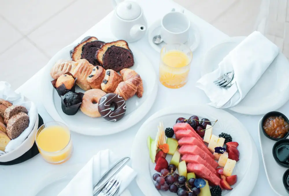 Desayuno variado con frutas, repostería, jugo de naranja y café sobre una mesa blanca.