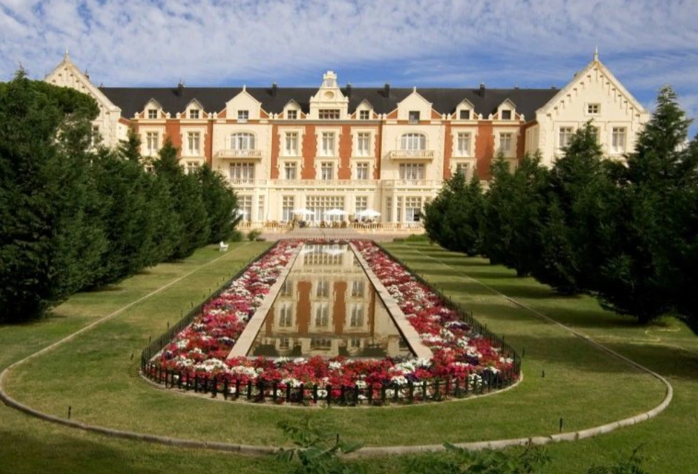 Gran edificio cl&aacute;sico con fachada elegante, jard&iacute;n sim&eacute;trico y fuente, rodeado de &aacute;rboles altos.