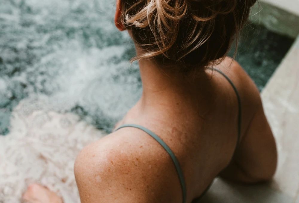 Mujer con cabello recogido vista de espaldas, sentada al borde de una piscina o jacuzzi.