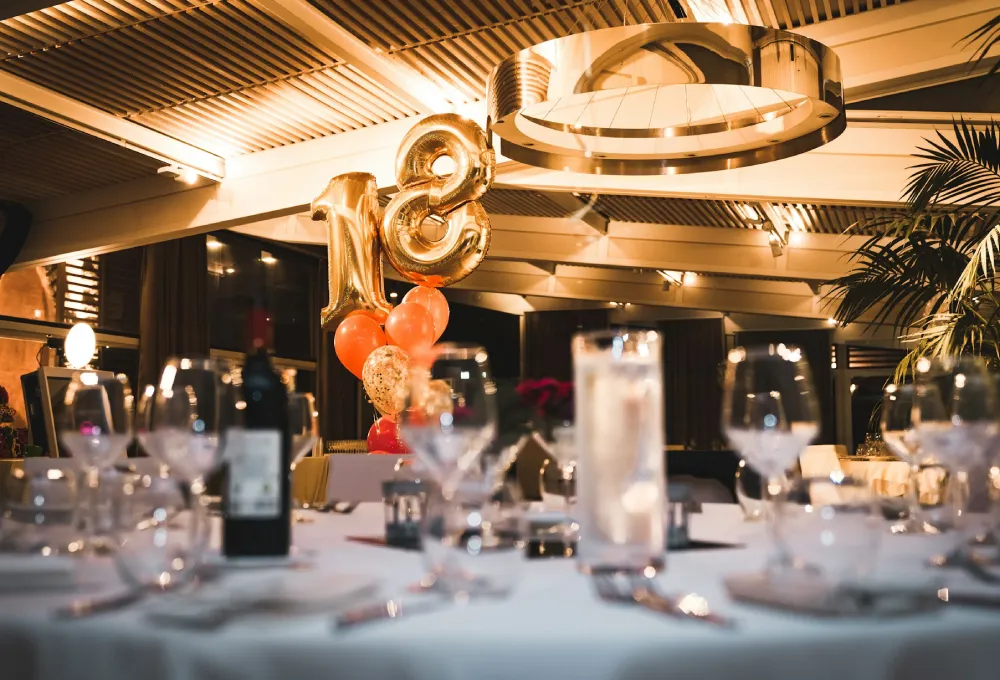 Mesa decorada para cumpleaños con globos dorados de número 18 y copas de vino vacías.