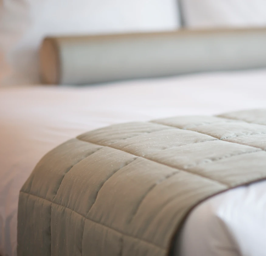 Cama con cobertor acolchado gris y cojín cilíndrico sobre sábanas blancas.