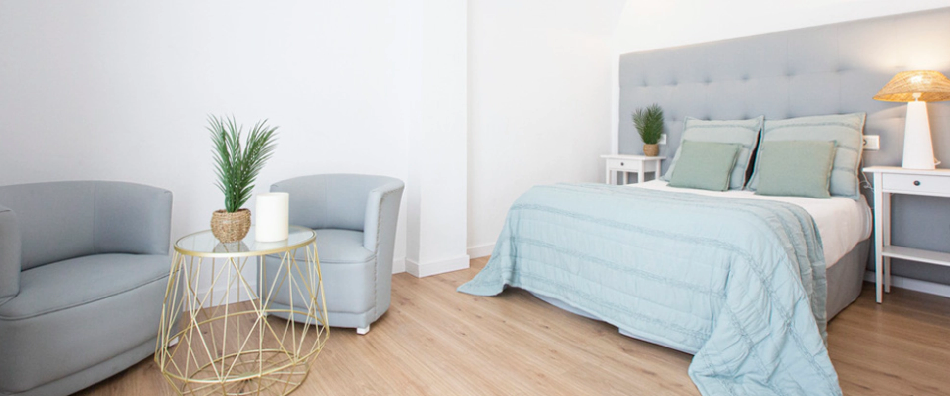 Habitación moderna con cama, sillones grises, mesa dorada y decoración minimalista.