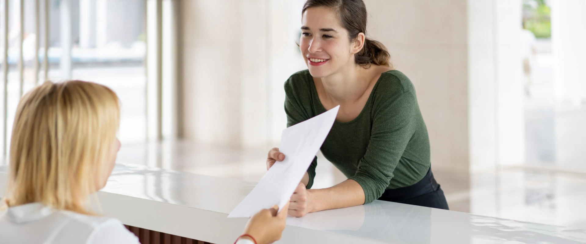 Mujer sonriendo y entregando documento a otra persona en un mostrador blanco.