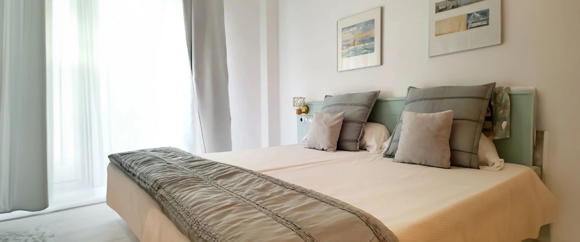 Cama doble con cojines grises, sábanas claras y cuadros en la pared. Cortina blanca a la izquierda.