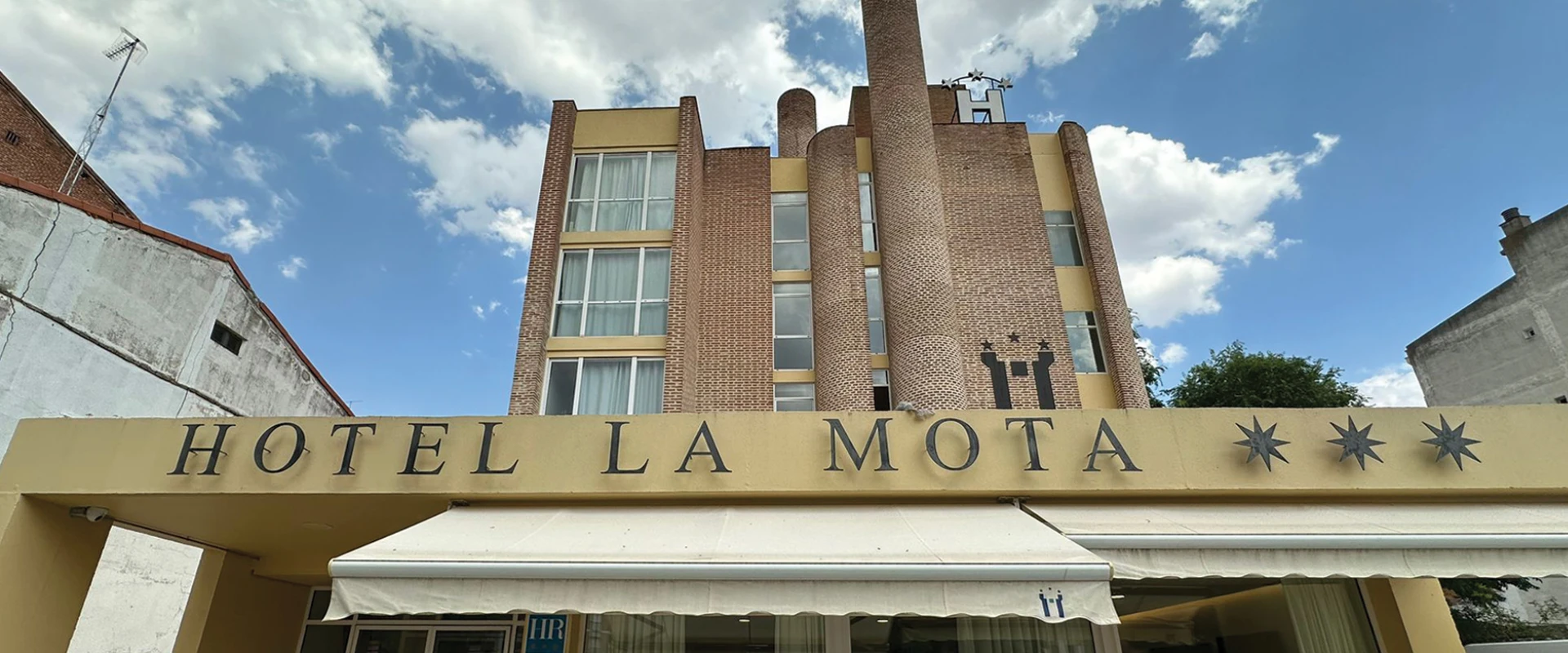 Fachada del Hotel La Mota con cielo azul y nubes de fondo.
