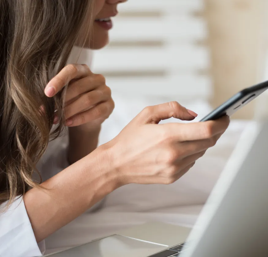 Persona en cama usando un teléfono móvil, con laptop enfrente y cabello castaño suelto.