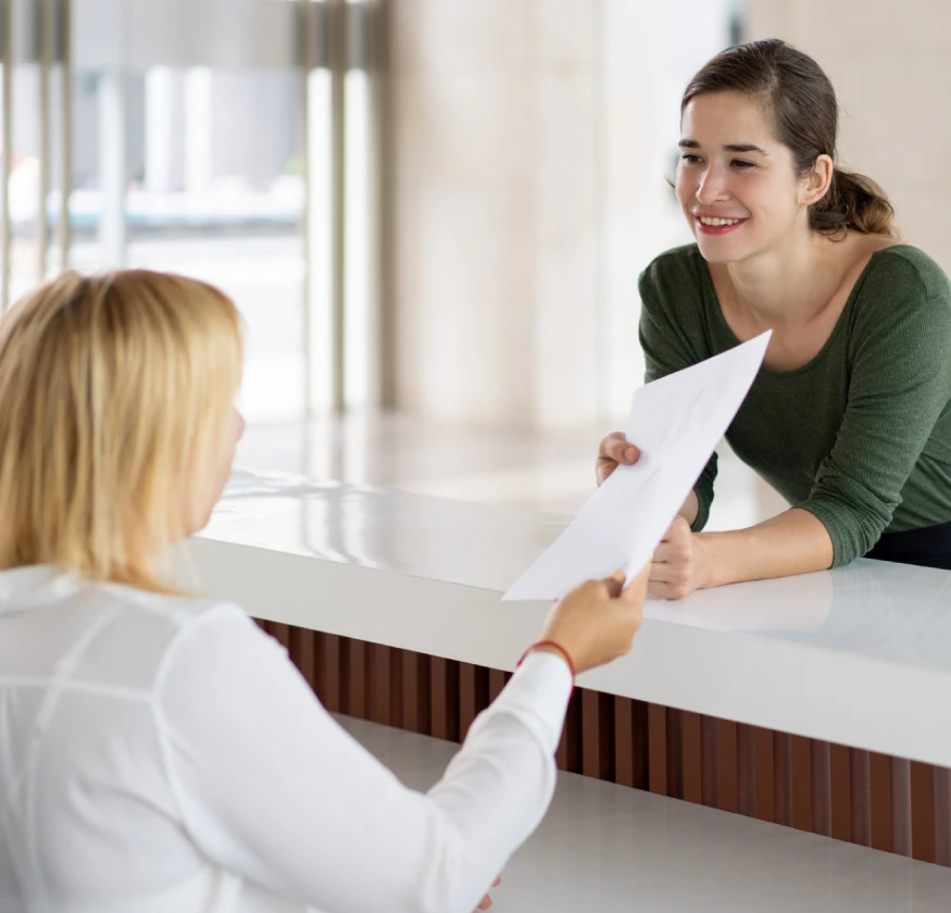Mujer en recepción entrega un documento a otra mujer sonriendo detrás del mostrador.