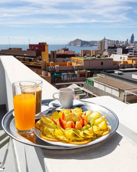 Desayuno en terraza con vista a la ciudad: plato de frutas, café y jugo de naranja.