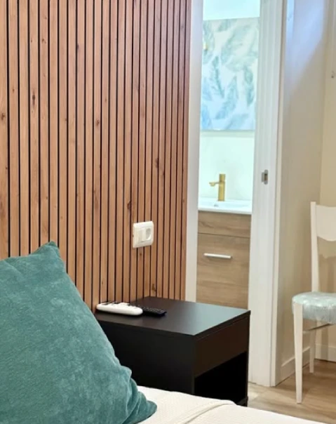 Dormitorio con pared de madera, cojín verde, mesita negra y vista de un baño al fondo.