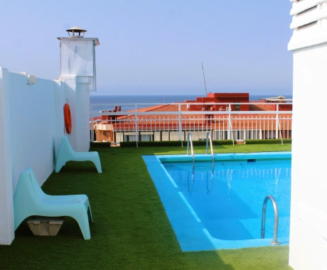 Piscina en la azotea con tumbonas, cerca del mar y edificios rojos al fondo.