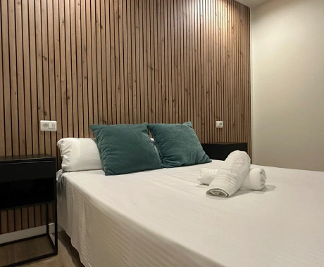 Cama con sábanas blancas, cojines verdes y toallas, con fondo de pared de listones de madera.