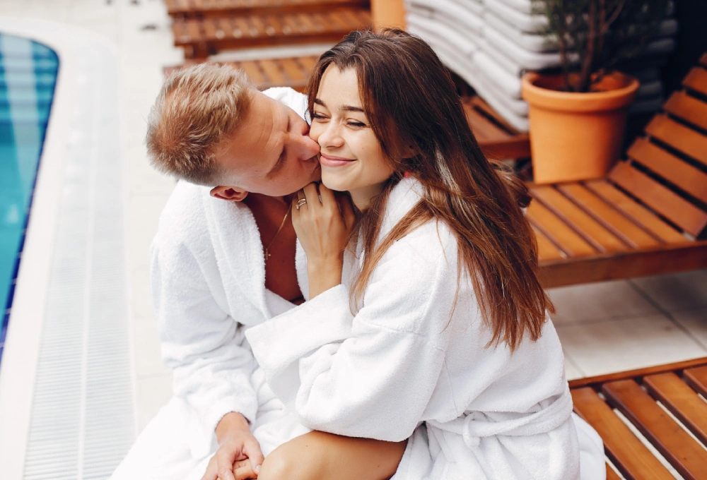 Pareja en batas blancas se abraza junto a una piscina, en un ambiente relajado y soleado.