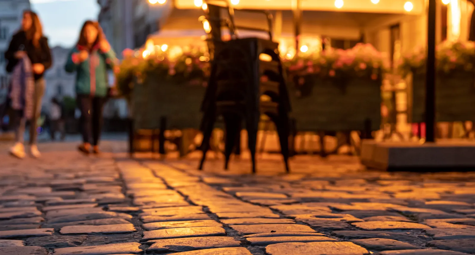 Calles de adoquines al atardecer con luces cálidas y personas desenfocadas al fondo.