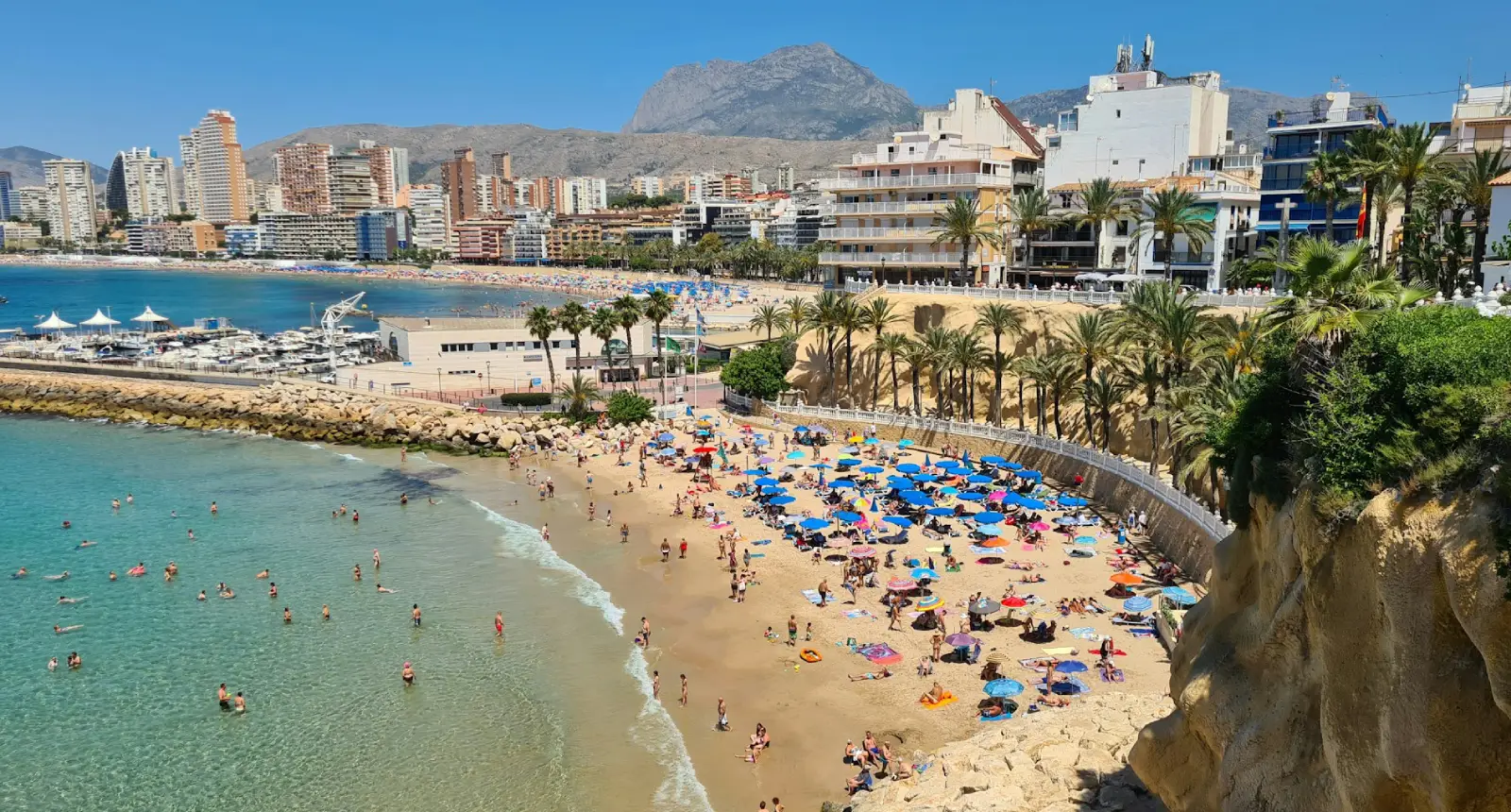 Playa con sombrillas, edificios y montañas al fondo, día soleado y muchas personas en el agua.