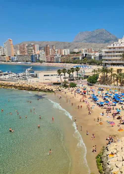Playa llena de bañistas y sombrillas, edificios y montañas al fondo bajo un cielo despejado.