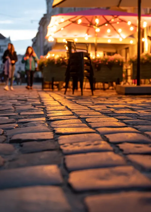 Calle empedrada al atardecer, con luces cálidas y dos personas caminando cerca de una terraza.