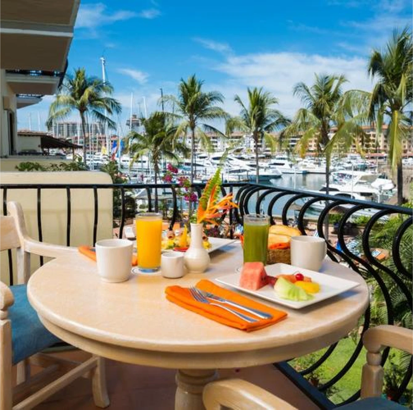 Desayuno en un balcón con vista al puerto y palmeras, incluye jugos, frutas y café sobre una mesa redonda.