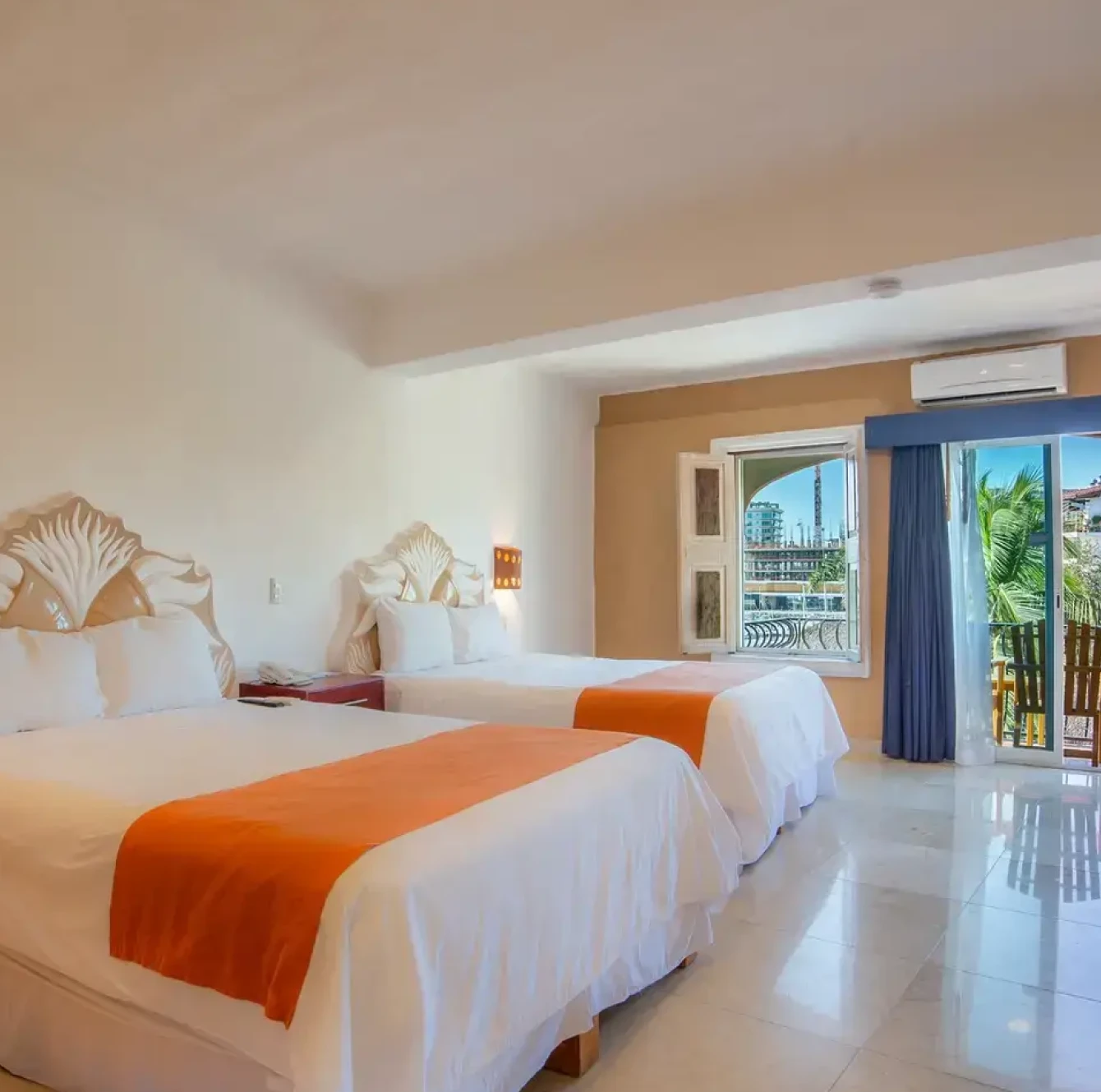 Habitación de hotel con dos camas, edredones blancos y cobertores naranjas. Ventana amplia con vista a la ciudad.