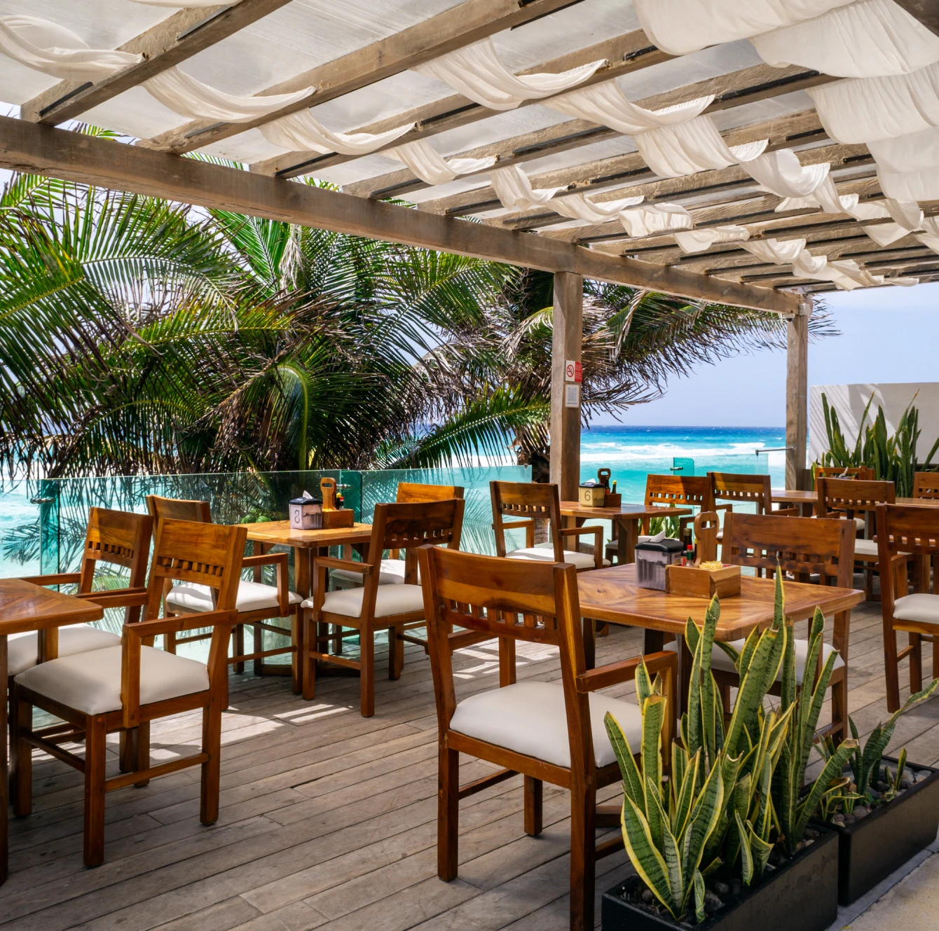 Terraza al aire libre con mesas y sillas de madera, frente al mar, con palmeras y cielo despejado.