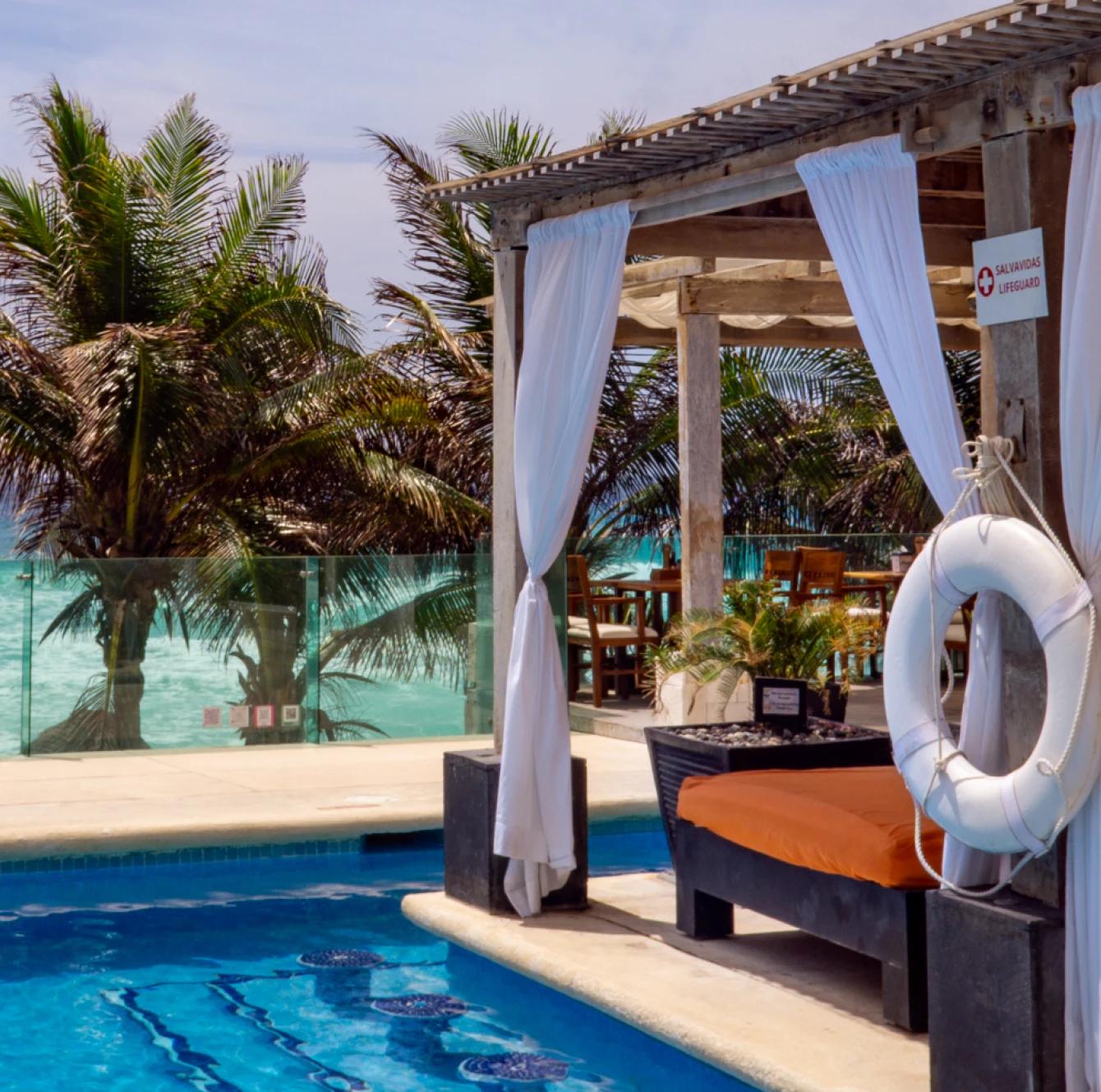 Tumbona con cortinas blancas junto a una piscina, con palmeras y el mar de fondo. Flotador colgado en la estructura.