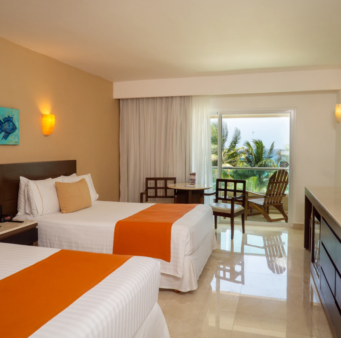 Habitación de hotel con dos camas, mantas naranjas, y balcón con vista a palmeras. Muebles de madera y piso de mármol.