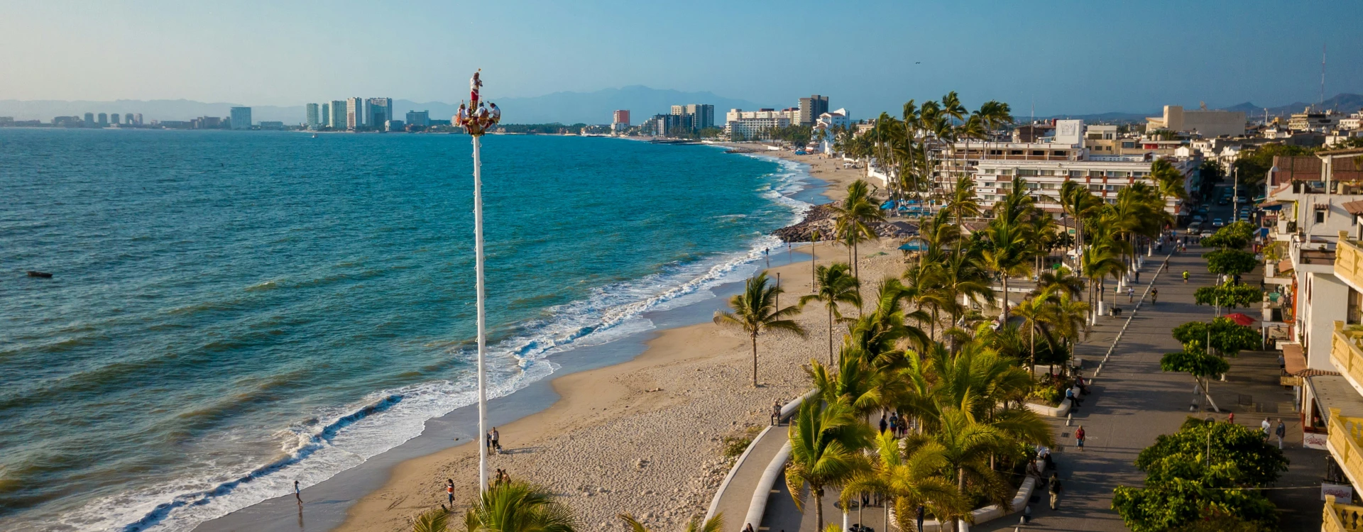 Vista aérea de una playa con un muelle, edificios al fondo y personas caminando por el malecón bordeado de palmeras.