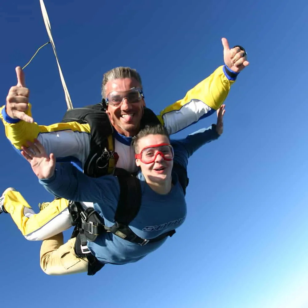 Dos personas haciendo paracaidismo, sonriendo bajo el cielo despejado.