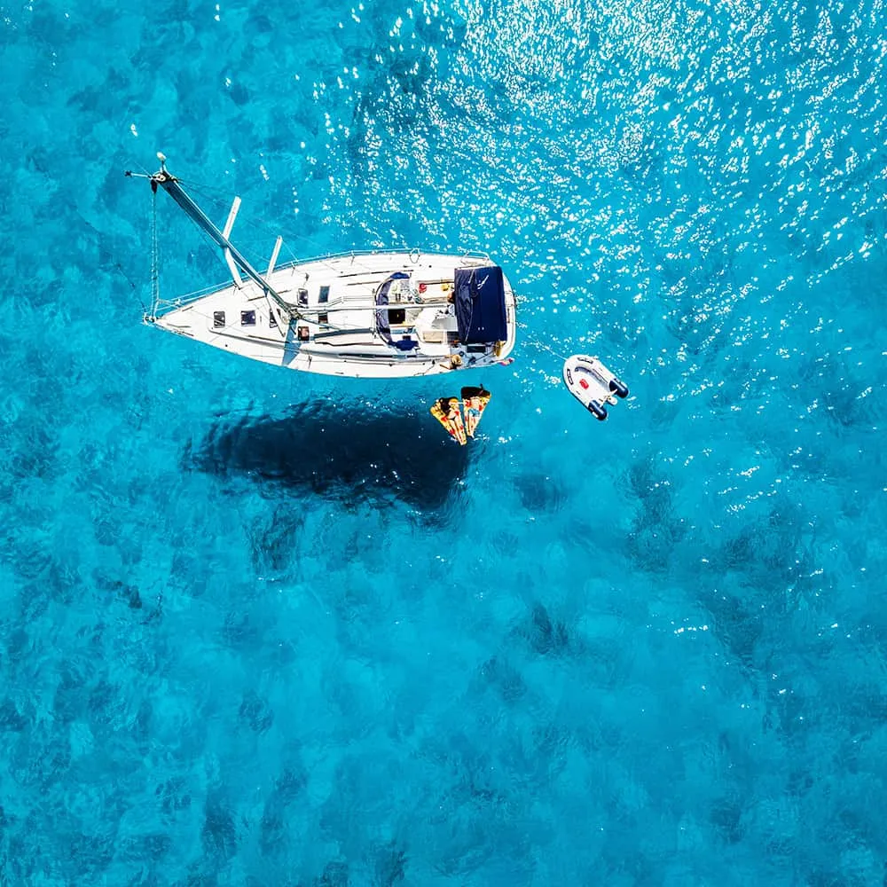 Velero blanco en agua cristalina con bote inflable cerca.