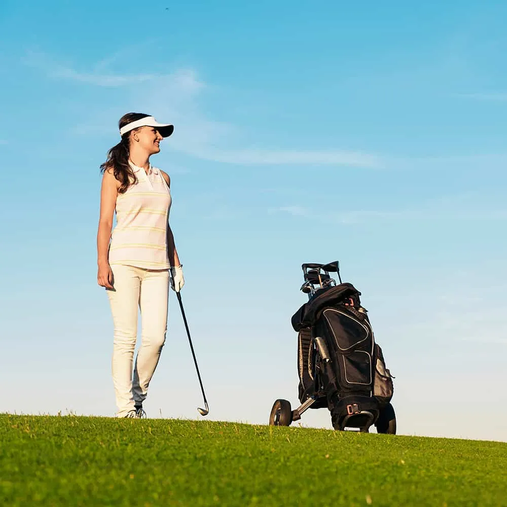 Mujer jugando golf en un campo con bolsa cerca, cielo despejado.