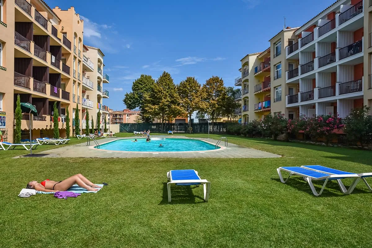 Piscina en un complejo de apartamentos con c&eacute;sped y tumbonas.