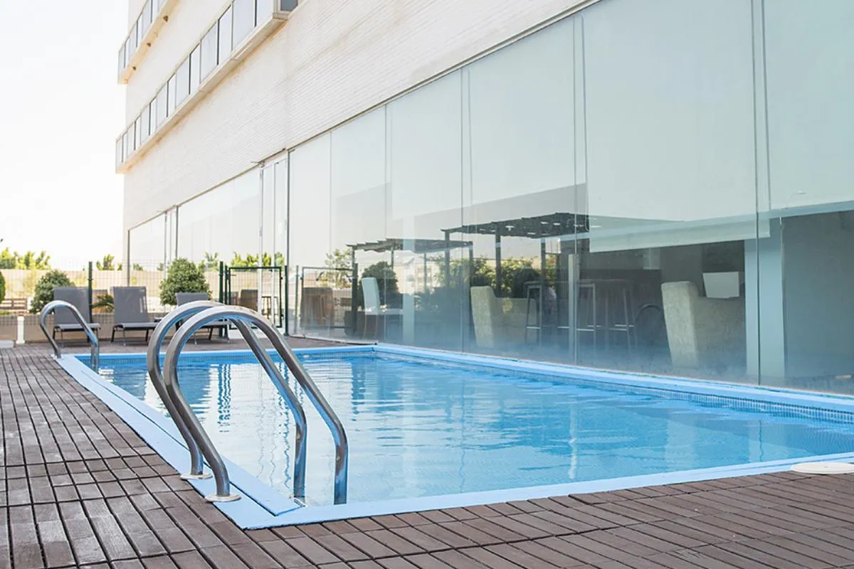 Piscina al aire libre junto a un edificio con pared de vidrio y suelo de madera.